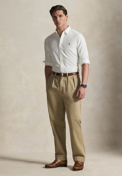 Nuori mies seisoo valkoinen pitkähihainen paita, beige pliseeratut housut, ruskea vyö, ruskeat kengät ja rannekello vasemmassa ranteessa.