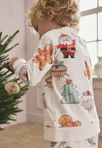 Light cream pajama set features festive patterns of Santa, gingerbread men, and Christmas trees. Soft cotton fabric with ribbed cuffs.