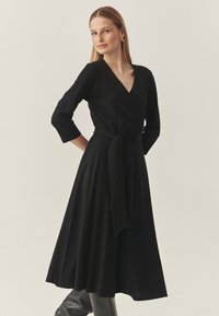 Zwarte wikkeljurk met lange mouwen, een V-hals, een getailleerde taille met een bijpassende riem en een uitlopende rok die tot halverwege de kuit reikt.