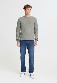 Siv zbican pulover s hribasto teksturo in okroglim izrezom, v kombinaciji z modrimi kavbojkami in belimi supergami s črnimi črtami, postavljenim pred svetlo ozadje.