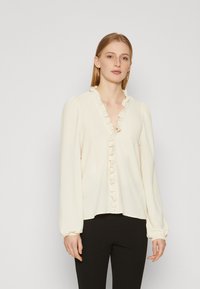 Cream-colored blouse with a ruffled v-neckline, long sleeves with gathered cuffs, and a relaxed fit. Worn over black slim-fit pants.