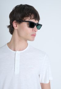 Occhiali da sole neri con design angolare e lenti verdi, indossati con una t-shirt bianca a maniche lunghe con scollatura a tre bottoni e logo ricamato.