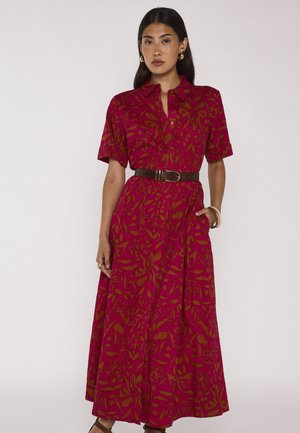 Donna che indossa un vestito lungo a maniche corte rosso e marrone con motivo, con cintura nera, orecchini dorati e braccialetti, in piedi davanti a uno sfondo semplice.