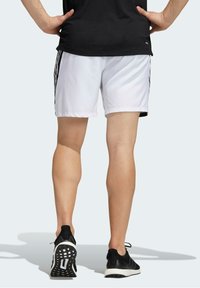 Valkoiset urheilushortsit mustilla sivuraitojen kanssa, yhdistettynä mustiin tennareihin, joissa on teksturoitu pohja ja valkoinen yksityiskohta. Takakatsaus.