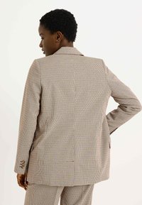 Blazer à carreaux en beige, marron et rouille. Présente une coupe ajustée, des revers crantés, deux poches latérales et des poignets à boutons. Tissu texturé.