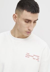 Sudadera blanca con cuello redondo, que presenta texto impreso en rojo en la zona izquierda del pecho. Textura suave y ajuste relajado.