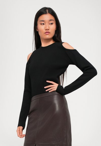 Woman with long dark hair wearing a black cold-shoulder top and a dark brown leather skirt, posing with one hand on her hip.