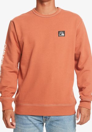 Homme portant un sweat-shirt à manches longues couleur rouille avec un petit écusson logo Quiksilver noir sur la poitrine et un texte blanc sur la manche gauche.