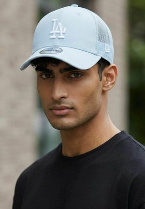 Giovane uomo con capelli corti e ricci che indossa un cappellino da baseball LA azzurro e una maglietta nera, guarda direttamente la fotocamera all'aperto.