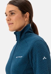 Giacca in pile blu con zip, caratterizzata da un motivo testurizzato, colletto alto e logo bianco sul petto e sulla manica. Materiale morbido con una vestibilità aderente.