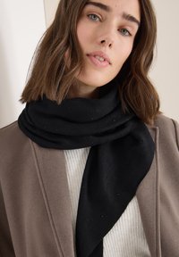 Bufanda negra con un ligero brillo envuelta alrededor del cuello, combinada con un abrigo beige y un suéter blanco texturizado, sobre un fondo liso.