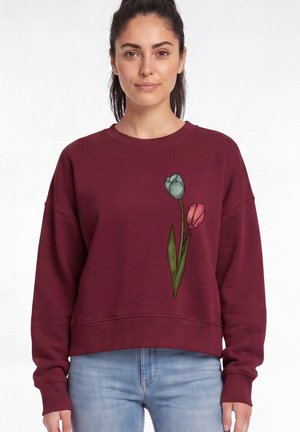 Femme portant un sweat-shirt bordeaux avec des motifs de tulipes bleues et roses à l'avant, associé à un jean bleu clair sur un fond uni.