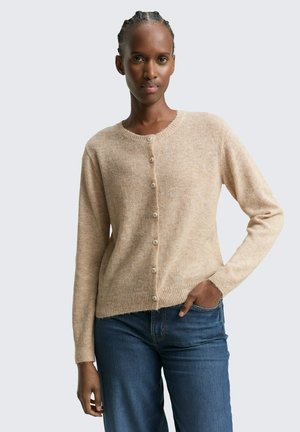 Cardigan en tricot beige en matériau doux, avec un col rond, une fermeture à boutons et des manches longues. Porté sur un jean bleu.