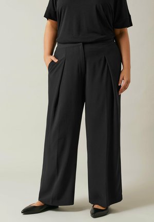 Black, wide-leg trousers with a pleated front and side pockets, made of a smooth fabric. Paired with a loose-fitting black top and flat shoes.