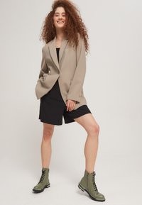 Blazer beige con spalle strutturate, pantaloni shorts neri e stivaletti alla caviglia verde con lacci. Tessuto testurizzato e vestibilità su misura.}}