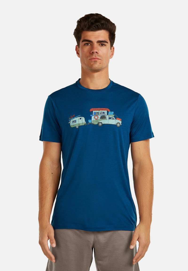 Mężczyzna w niebieskim t-shircie z grafiką samochodu holującego kampera z rzeczami na zewnątrz, stojący na tle jednolitego tła.