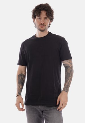 Hombre con cabello rizado y tatuajes en ambos antebrazos, vistiendo una camiseta negra y vaqueros grises, de pie frente a un fondo claro y liso.