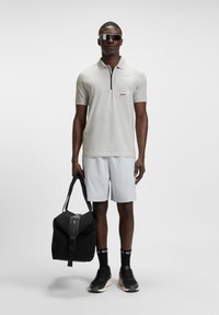 Man wearing light gray polo shirt and shorts, black sneakers and socks, dark sunglasses, holding black duffel bag with strap.