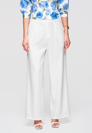 Femme portant un pantalon blanc large taille haute, une chemise boutonnée bleue à fleurs, des sandales blanches à talons, et un bracelet en or au poignet gauche.