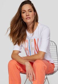 White short-sleeve t-shirt featuring colorful stitched lettering "Tout va BIEN." Model sits on a wire chair, wearing coral trousers.