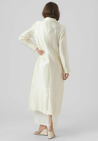 Lång, elfenbensfärgad kappa i texturerad tyg med krage, långa ärmar och sidoklyftor, parat med en vit lager-på-lager klänning och nakna kilklackar.