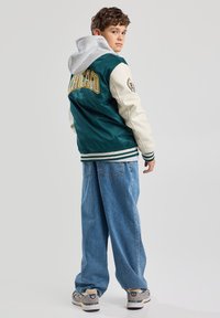 Varsity jacket in teal and cream with "CHICAGO" lettering, gray hood, ribbed cuffs, and loose blue denim pants. Grey sneakers with accents.