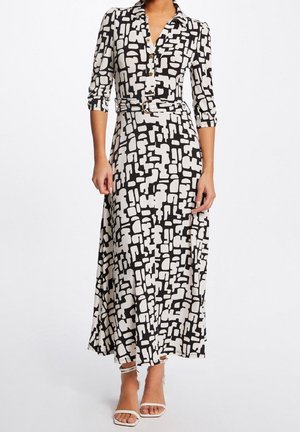 Kobieta ubrana w długą sukienkę z czarno-białym abstrakcyjnym wzorem, z paskiem, rękawami do połowy przedramienia oraz białe szpilki z paskiem wokół kostki.