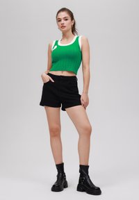 Roheline soonikcrop top valgete detailidega, mustad denim-lühikesed püksid ja mustad pahkluu saapad, millel on pandlad. Neutraalne taust.