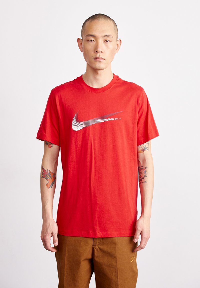 Red cotton t-shirt featuring a large white Nike logo with linear detail across the chest. Model wears brown pants and has tattoos visible.