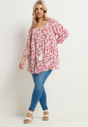 Frau in einer weißen Bluse mit roten Blumen, blauen Skinny-Jeans und weißen Sandalen, die vor einem einfarbigen Hintergrund steht.