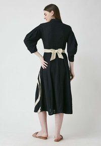 Schwarzes Baumwollkleid mit Dreiviertelärmel, einer taillierten Taille, gebunden mit einem cremefarbenen Band, und einem fließenden Rock mit einem seitlichen cremefarbenen Streifen.