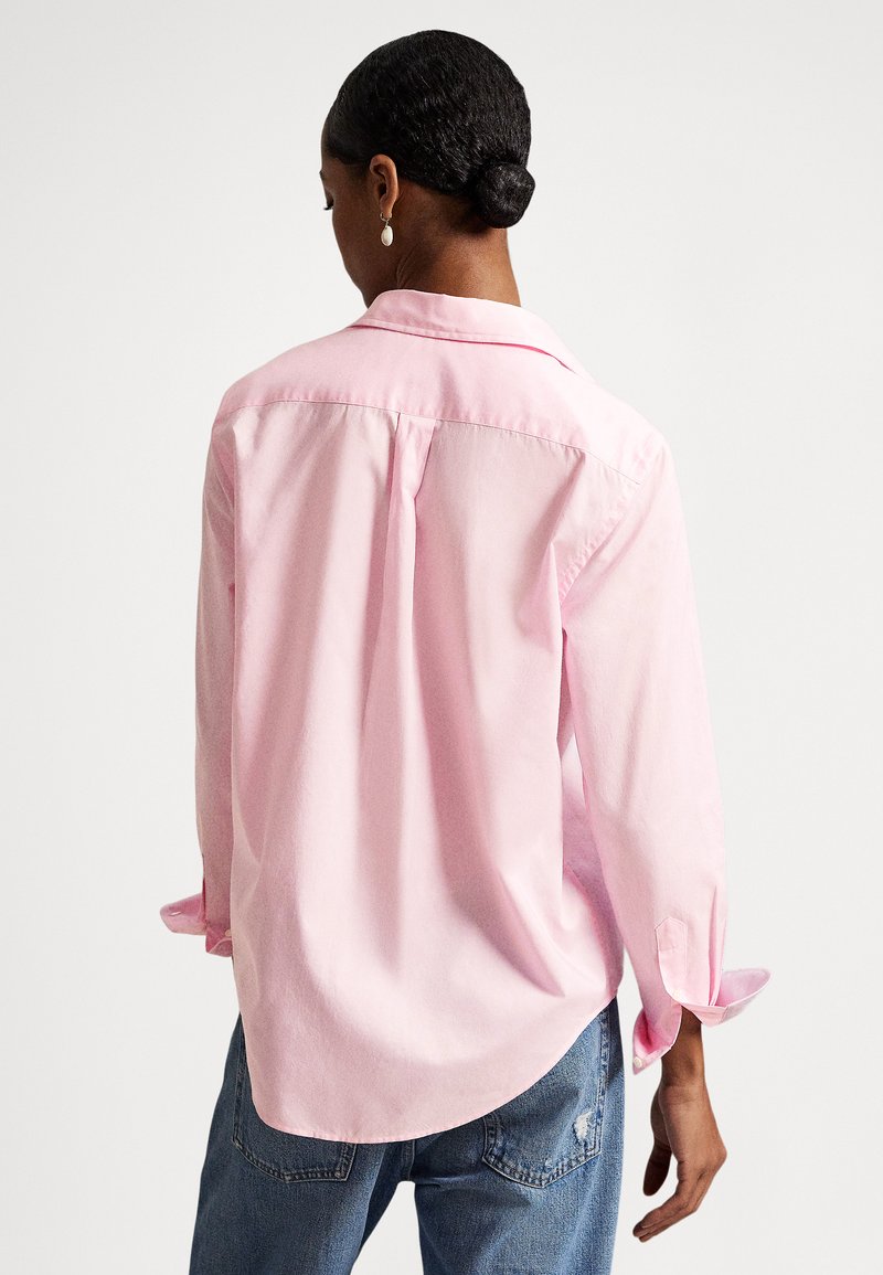 Donna con capelli scuri raccolti in uno chignon basso, indossa una maglia a maniche lunghe rosa chiaro e jeans blu, vista di spalle su uno sfondo bianco semplice.