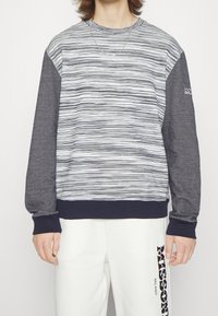 Sudadera de cuello redondo con rayas horizontales grises y blancas, mangas gris oscuro, puños y bajo acanalados. La parte delantera presenta un sutil detalle de logo.