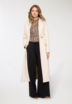 Cappotto beige lungo con due bottoni, indossato sopra una maglia con stampa leopardata e pantaloni neri a gamba larga. Tessuto liscio con design strutturato.