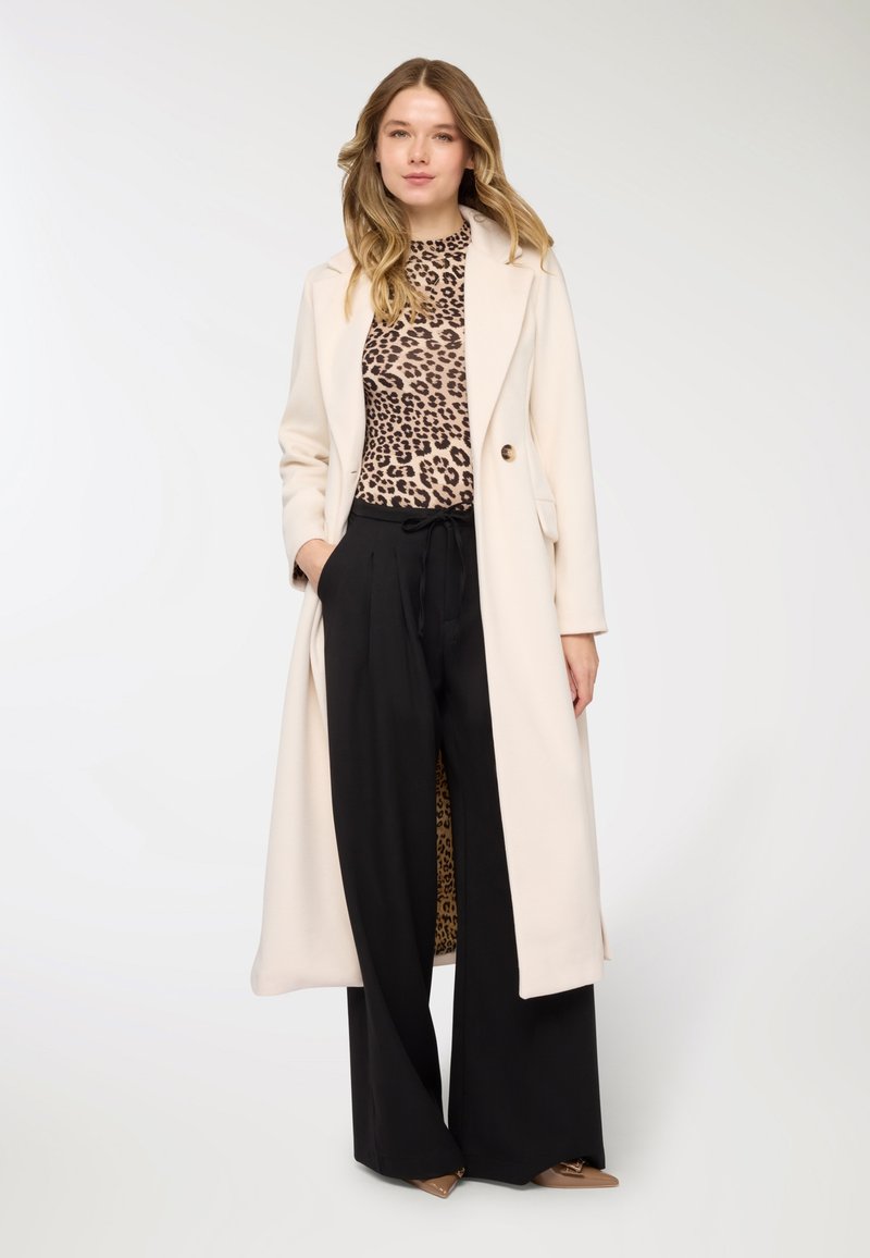 Cappotto beige lungo con due bottoni, indossato sopra una maglia con stampa leopardata e pantaloni neri a gamba larga. Tessuto liscio con design strutturato.