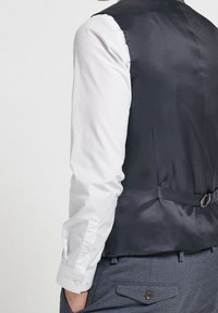 Marineblaues Satin-Weste mit einem eleganten Design, das eine Rückensteckschließe und eine glatte Textur aufweist, kombiniert mit einem weißen Hemd und grauen Hosen.