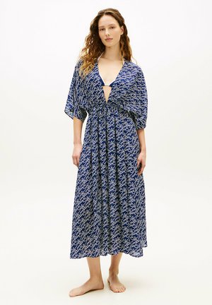 Kvinde med krøllet hår iført en blå mønstret midi-kjole med brede ærmer og dyb V-udskæring, stående barfodet på en hvid baggrund.