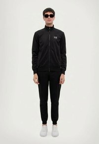 Άνδρας φορώντας μαύρο tracksuit EA7, μαύρα γυαλιά ηλίου και λευκά παπούτσια στέκεται σε σκηνικό με απλό φόντο.