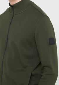 Olivově zelená bunda na zip s vysokým límcem, žebrovanou strukturou a černou nášivkou na rukávu; látka působí měkce a pružně.