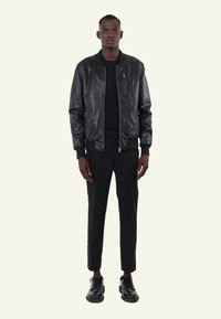 Zwarte leren bomberjack met ribgebreide manchetten en een ritszak, gedragen over een zwarte ronde hals t-shirt en zwarte tapered broek, afgewerkt met zwarte schoenen.