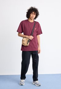 Burgundy short-sleeve t-shirt, black pants, and gray sneakers. A tan crossbody bag with zippers is worn on the shoulder. Plain background.
