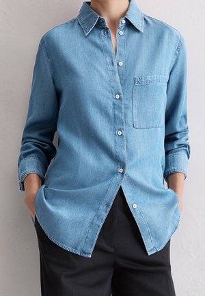 Persoon die een lichtblauw denim hemd met knopen en een borstzak draagt en een zwarte broek, handen in de zakken, staand tegen een effen muur.