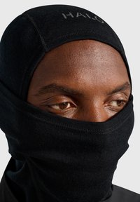 Svart balaklava tillverkad av elastiskt tyg, med slät textur, figursydd design och subtil branding i pannan.