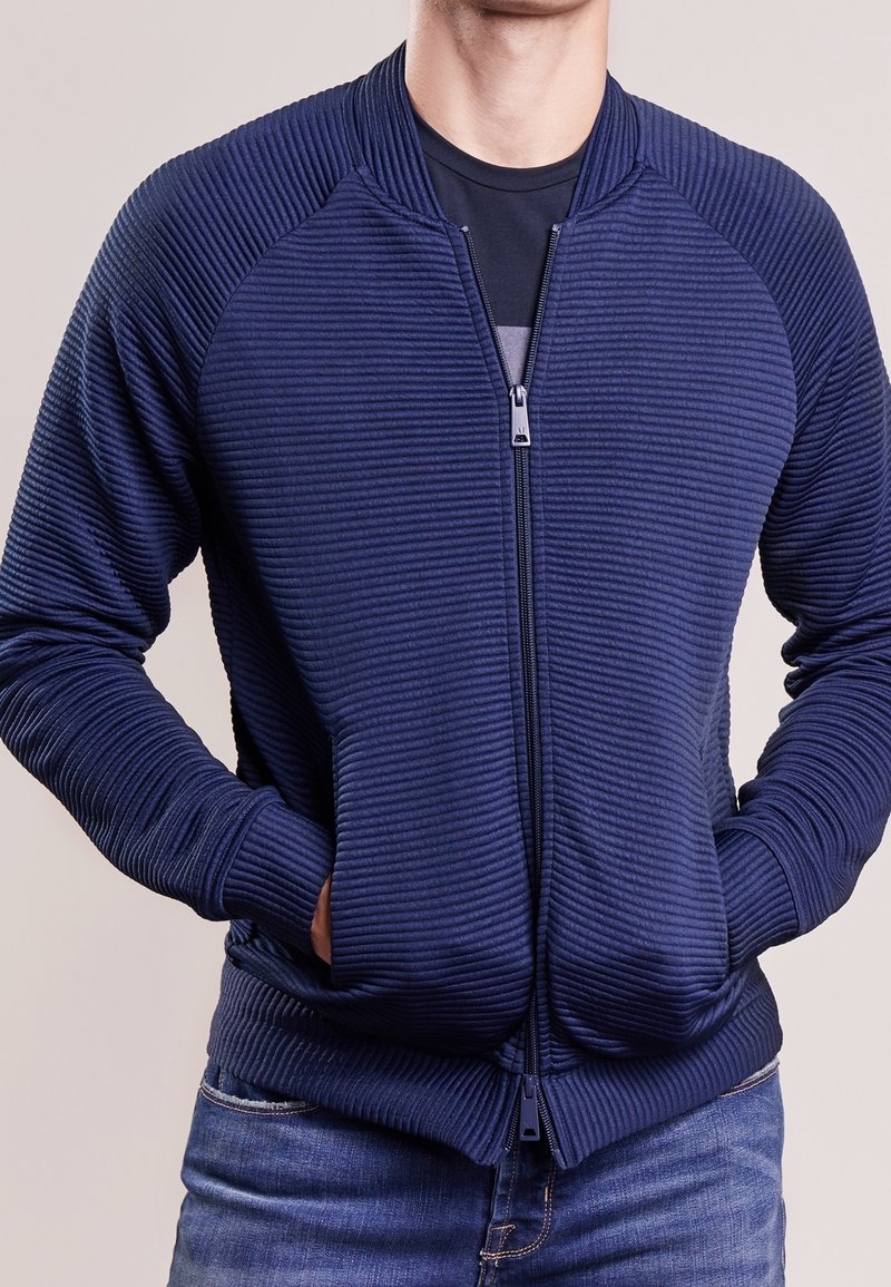 Homme portant une veste zippée côtelée bleu marine par-dessus un t-shirt noir, les mains dans les poches de la veste, assortie à un jean bleu.