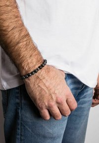 Brățară din mărgeluțe negre, cu mărgele netede și rotunde, purtată pe un încheietura mâinii, asortată cu un tricou alb și blugi, evidențiind o suprafață texturată.
