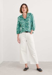 Grüne Bluse mit floralen Motiven und Dreiviertelärmeln, kombiniert mit weit geschnittenen weißen Hosen und schwarzen Clogs. Verfügt über einen lässigen Schnitt und einen Knopfverschluss.