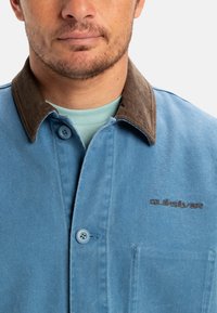 Light blue jacket with a brown corduroy collar, featuring a single button closure, chest pocket, and embroidered logo. Smooth fabric texture.
