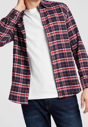 Mężczyzna w odpiętej czerwono-biało-niebieskiej koszuli w kratę na białym t-shircie i ciemnoniebieskich dżinsach, ręka przy szyi.