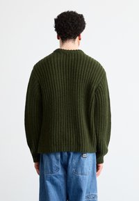 Ελαιόχρωμο πλεκτό πουλόβερ με υφή ριμπ, στρογγυλή λαιμόκοψη και άνετη εφαρμογή, συνδυασμένο με μπλε τζιν με πίσω τσέπες.
