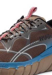 Gros plan d'une basket HUGO marron et bleue avec des lacets noirs, des empiècements gris et une languette à tirette rouge et blanche.